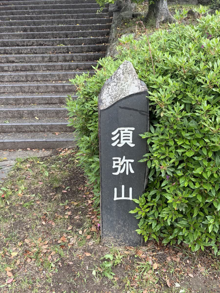名古山霊苑の須弥山と書かれた黒い石碑、緑の生垣と石段の前に立つ