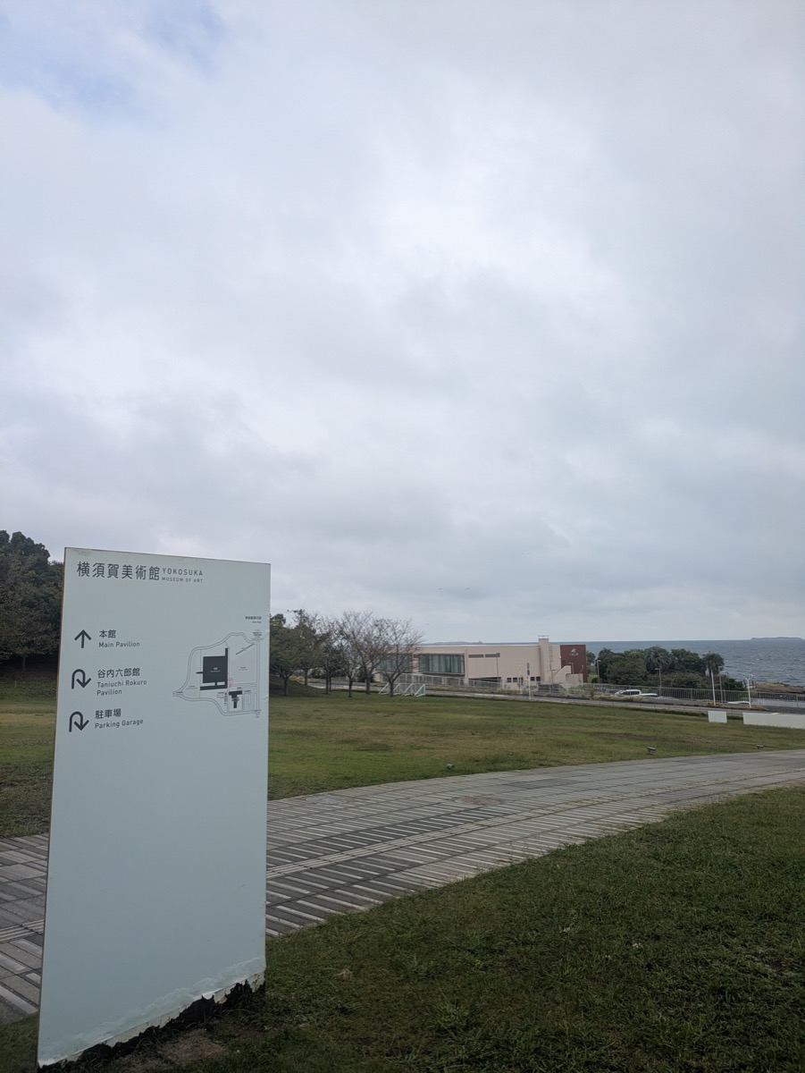 横須賀美術館の芝生広場に立つ案内サイン。奥に低く横長の美術館と海が見える