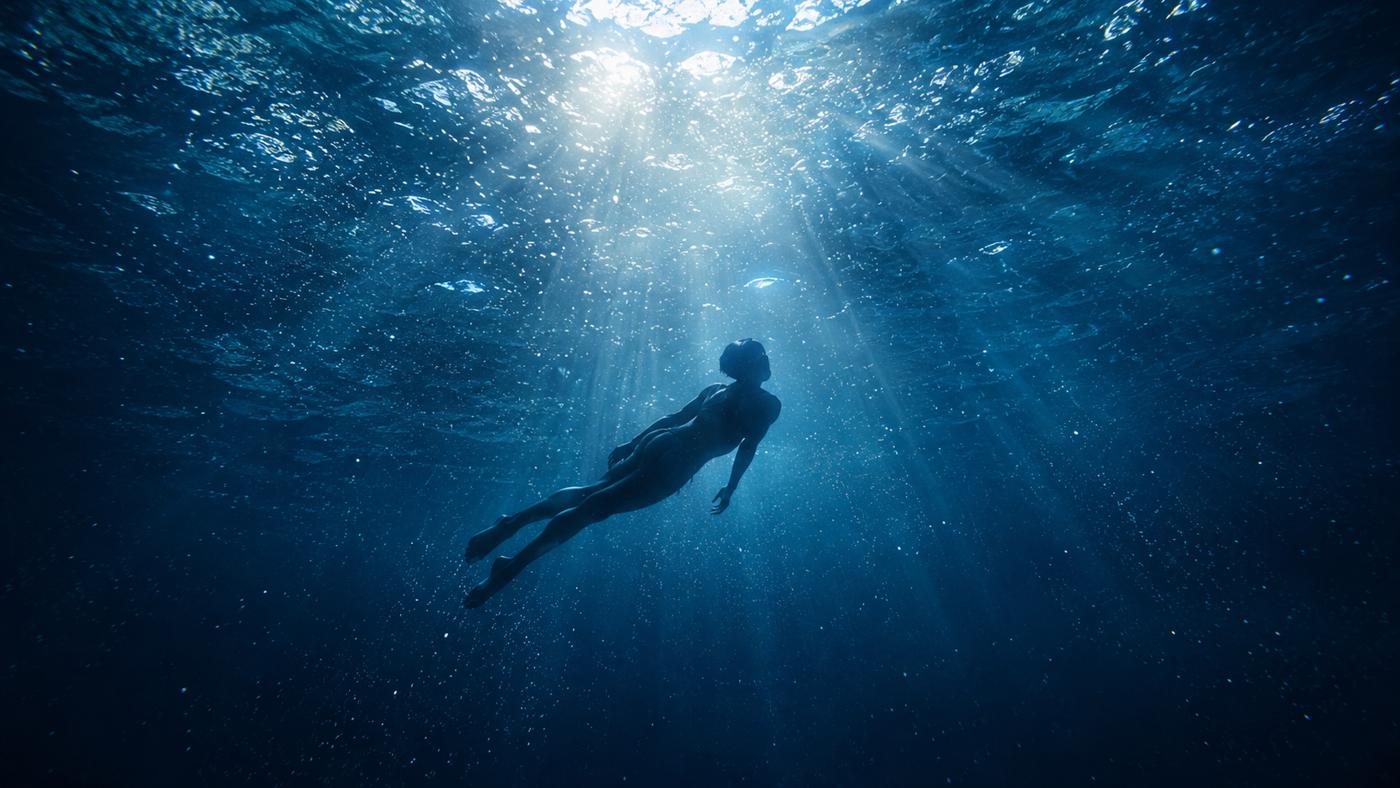 青い水中を静かに泳ぐスイマー。水面から光のカーテンが差し込み、宇宙空間のような静けさ