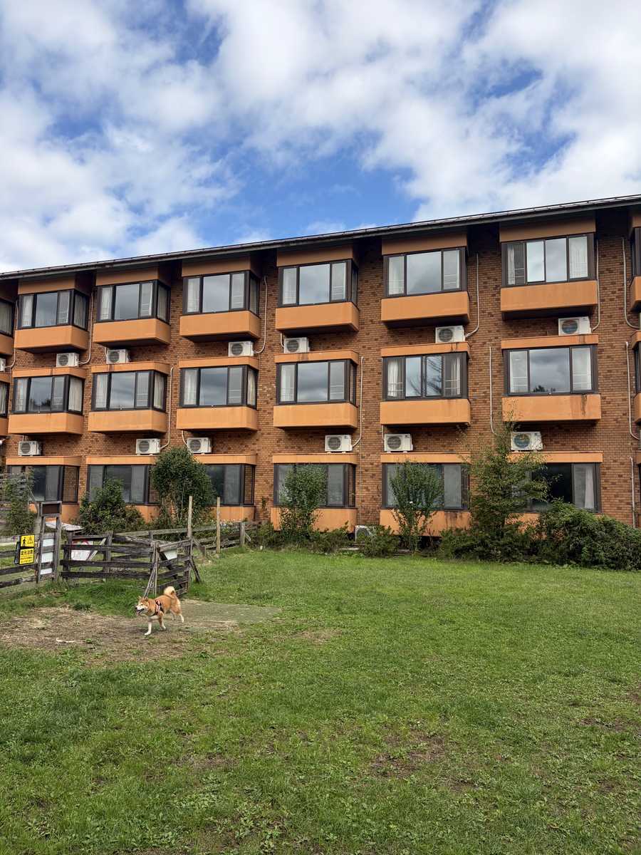 青空の下、芝生の向こうに建つ3階建てのレンガ色のホテル棟。手前の芝生に柴犬もみじが小さく写る